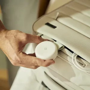 Twelve South Butterfly™ SE kompaktiškas baltas 2 viename Qi2 įkroviklis, laikomas rankoje šalia kelioninio krepšio