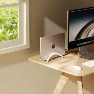 Twelve South BookArc Flex vertical laptop stand holding a MacBook upright beside a desktop monitor.