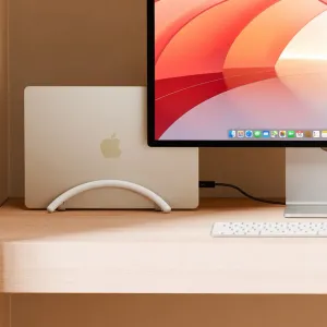 Twelve South BookArc Flex vertical laptop stand holding a MacBook upright beside a desktop monitor.