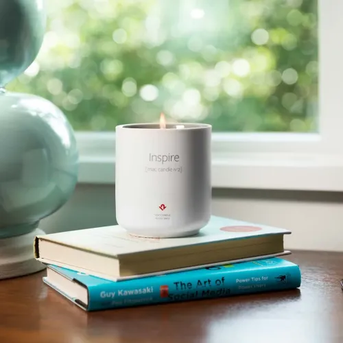 "Inspire Mac Candle No. 2" žvakė baltame cilindro formos keraminiame puodelyje su degančia dagtimi, padėta ant dviejų knygų ant stalo prie lango.