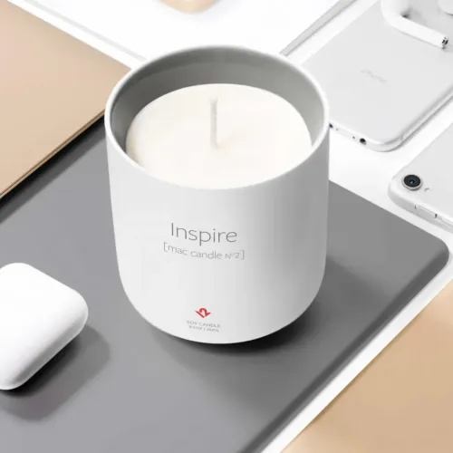 Hvitt sylindrisk keramikklys med teksten Inspire [mac candle n°2], åpen veke, på en grå plate ved siden av flere lyse tekniske duppeditter.