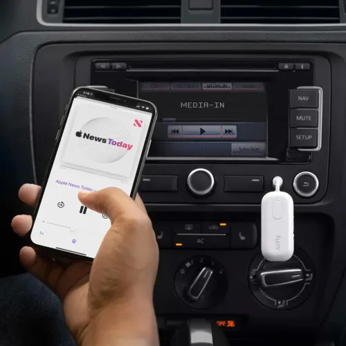 AirFly Deluxe, valkoinen sovitin, jossa on soikea painike ja LED, kytketty auton kojelautaan puhelinta pitelevän käden viereen.