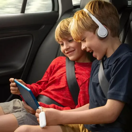 AirFly Deluxe valkoinen soikea sovitin lyhyellä 3,5 mm:n kaapelilla liitettynä tablettiin auton takapenkillä kahden pienen lapsen vieressä