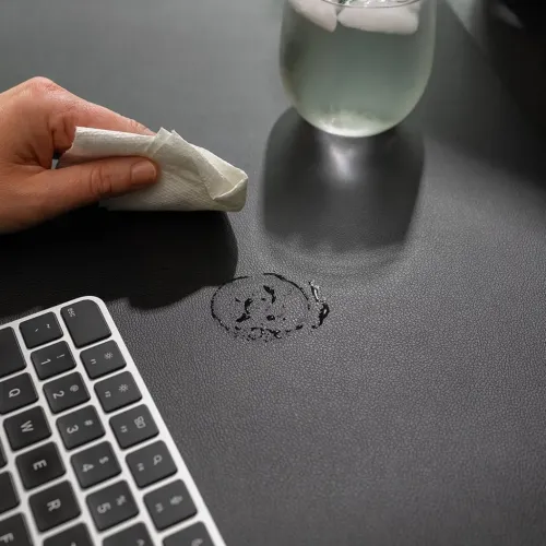 Twelve South DeskPad i sort tørres af med serviet; vand drypper ved siden af glas med is, tastaturet er synligt i nederste hjørne.