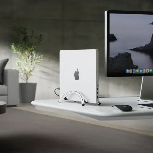 BookArc Flex-stativ i krom holder en MacBook vertikalt på et skrivebord; bueformet holder med føtter og synlige sideporter.