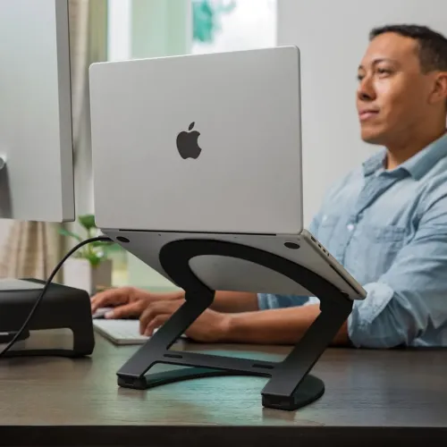 Black adjustable MacBook stand lifts a laptop onto desk; two articulated arms, curved base, side ports and cable visible.