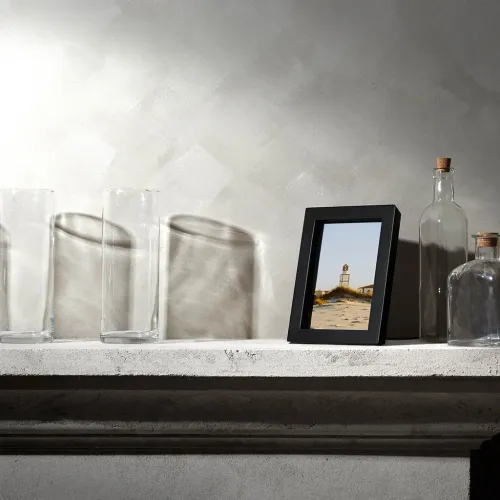 PowerPic-fotorammen i svart står i stående modus på en lys betonghylle, omgitt av glassflasker og med et strandmotiv i rammen.