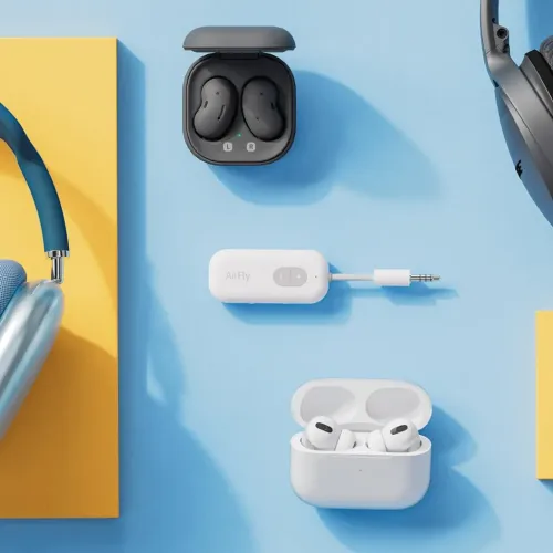 Twelve South AirFly SE, hvit adapter med 3,5 mm plugg og knapper, liggende på blå flate omgitt av hodetelefoner og ørepropper.