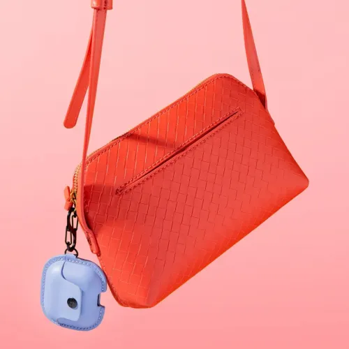 Läderfodral för AirPods i ljuslila med synlig knapp och svart S-klips, fäst vid röd vävd väska och hängd mot rosa bakgrund.