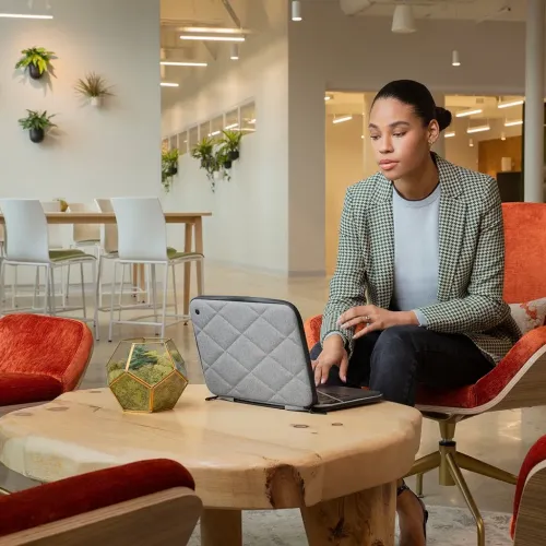Harmaa tikattu MacBook-kotelo Twelve South SuitCase, joka on kääritty tietokoneen ympärille mustalla vetoketjulla ja pyöristetyillä kulmilla pöydällä.
