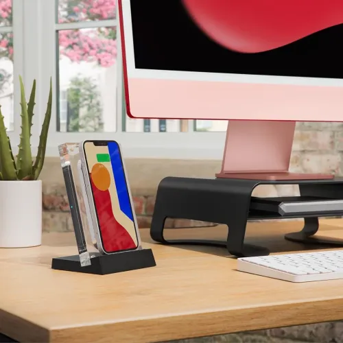 Trådløs laderamme i klar akryl med svart sokkel, vist i sideprofil på skrivebordet med telefonen stående mot rammen ved skjermen.