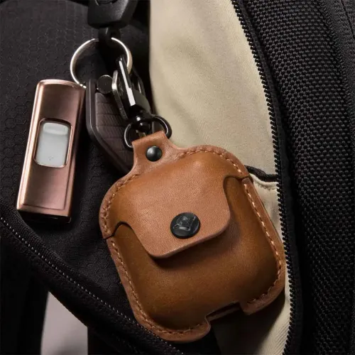 AirSnap-nahkakotelo AirPodsille konjakinvärisenä, jossa on nappisuljenta ja pohja-aukko, kiinnitetään klipsillä reppuun avaimenperässä.