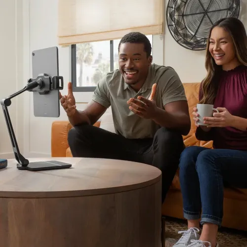 Svart justerbart iPad-ställ med ledad arm på bordsfot, håller surfplatta horisontellt på soffbord i vardagsrum med personer.