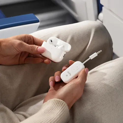 AirFly Pro 2 hvid lydadapter med 3,5 mm stik og knapper holdes i hånden ved siden af den åbne kasse på flysædet i skødet om bord.