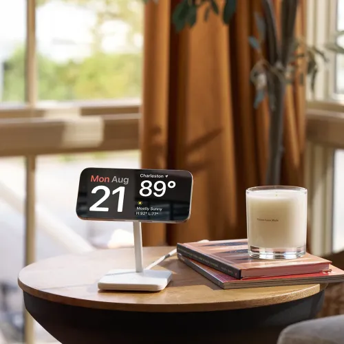 Vitt laddningsställ med vinklad stav och rektangulär bas håller en liggande iPhone på ett sidobord bredvid böcker och ljus.