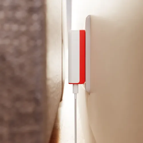 PlugBug 120W USB-C‑laddare i vitt och rött sitter platt i vägguttag bakom möbel, med smal profil och kabel nedåt intill.