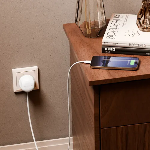 Hvit, flettet skjøteledning med rundt støpsel i EU-stikkontakt, koblet til en telefon på et nattbord av tre ved siden av.