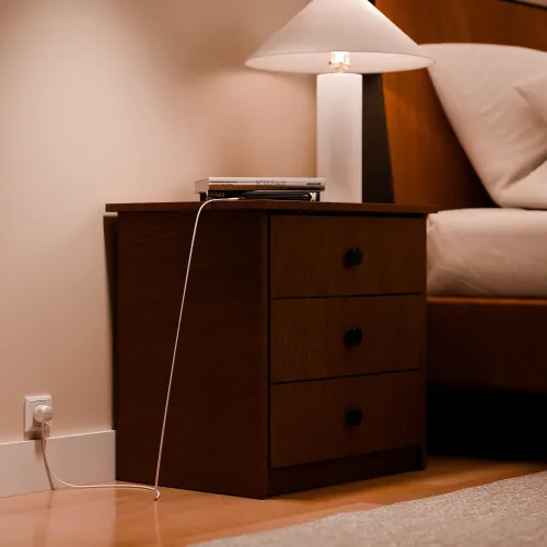 PowerCord vit förlängningssladd i EU-uttag; kabeln löper från vägguttaget upp längs ett nattduksbord i trä till ovansidan.