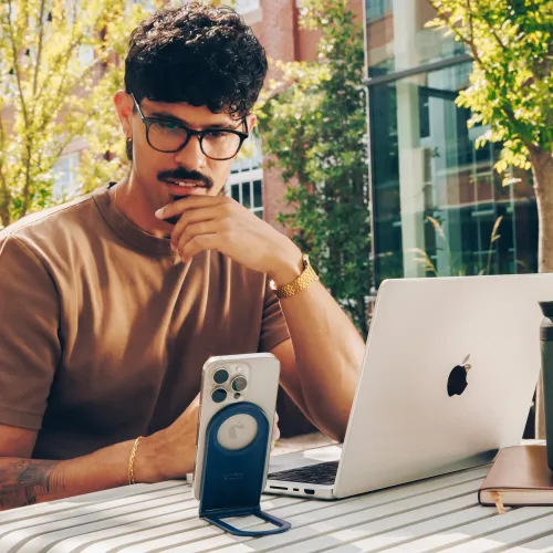 Curve Nano-telefonstativ i blått med magnetisk plate, utfellbart ben, holder telefonen vertikalt på et utendørs bord ved siden av den bærbare datamaskinen.